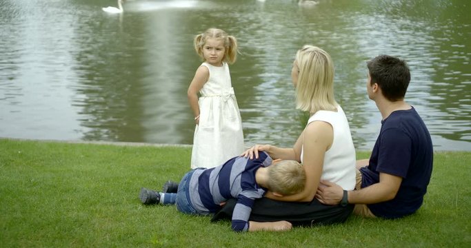 a young family with two children resting on the lawn near the lake. mother stroking her son on the head, daughter says something to parents and laughs