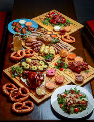 Flat-lay of Oktoberfest dinner table with grilled meat sausages, pretzel pastry, potatoes, cucumber salad, sauces, beers