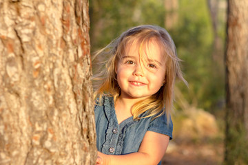 A cute blonde little girl playing hide and seek behind a tree at sunset