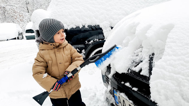 Close up photo of adorable little boy in beige coat helping to clean snow covered car with blue brush