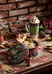 homemade delicious gingerbread cookies in vintage gift round metal box with christmas ornaments on wooden rustic red table with cup of coffee with foam, christmas tree branches opposite brick wall
