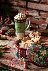 homemade delicious gingerbread cookies in vintage gift round metal box with christmas ornaments on wooden rustic red table with cup of coffee with foam, christmas tree branches opposite brick wall