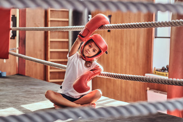 Beautiful kid girl is sitting on boxing ring wearing boxer uniform - gloves and helmet.