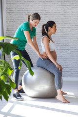 Physiotherapist helping to beautiful pregnant woman for doing pilates exercises with ball preparing...