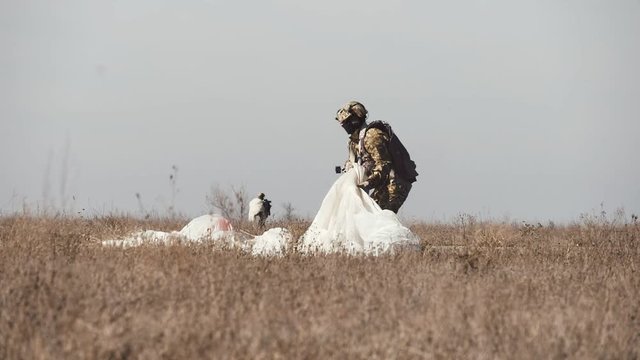 Military parachutist gathering his white parachute in a rough field in slo-mo 