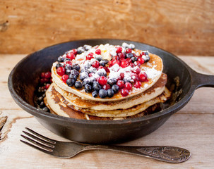Stack of homemade shrovetide carnival clean eating oat pancakes with powered sugar, blueberries and cowberries in cast iron pan on rustic country table with old silver dishware. Soft focus. Copy space