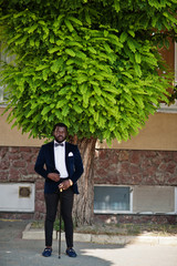 Handsome fashionable african american man in formal wear and bow tie with walking stick stay under...