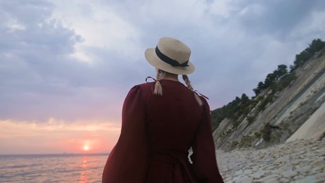 The view from the back. Slow motion Portrait of a young girl in a red dress and a straw hat on the seashore. A girl in the wind holds a hat with her hands. Waiting for the return of the sailors