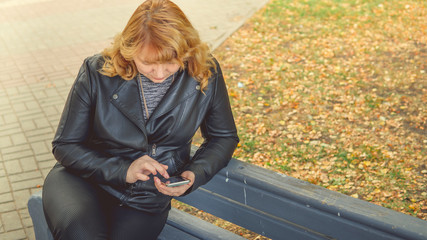 a grown woman sitting on bench in autumn Park dials the number on mobile phone