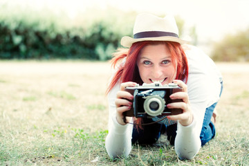 Pretty smiling redhead at ground taking pictures with an old camera. vintage treatment