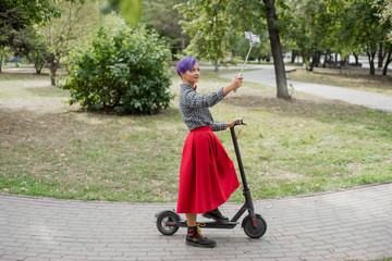 A young woman with purple hair rides an electric scooter in a park. Stylish girl with a shaved...