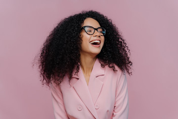 Joyous overjoyed dark skinned female manager has frizzy curly hair, laughs with happiness, wears...