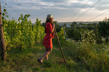 Beautiful teen girl in red dress and hat leaned on the rakes, resting while working in the vineyard, people and garden concept