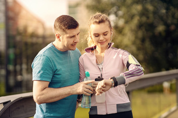 Smiling couple checking time or pulse on heart rate monitor watch - Runners in the park - Man and woman training outside in nature