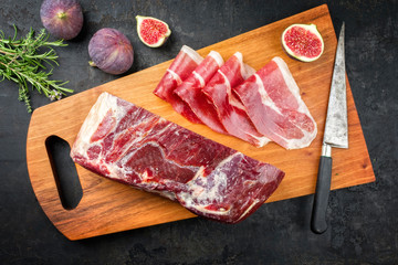 Traditional dry cured and smoked ham with a bouquet garni  and a ripe fig offered as top view on a modern design cutting board - obrazy, fototapety, plakaty