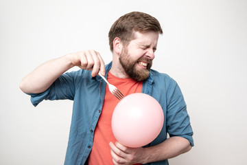 Bearded man is about to burst a balloon with a fork and bared his teeth in fear. Fun or stress...