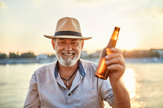 Portrait of a happy senior man drinking a beer at the riverside at sunset