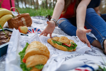 Picnic lunch outdoor in park on sunny day with croissants and sandwich. picnic on the nature with delicious fast food mael. woman hands reach for croissants in the park