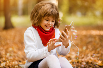 Outdoor portrait of little girl playing with phone. Happy child girl using smartphone in autumn park