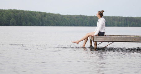 Woman sits on a pier and splashes in the lake with her legs. Young female model resting outdoors.