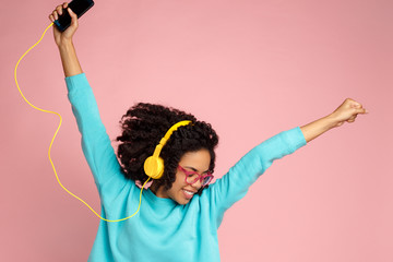Pretty african american young woman with bright smile dressed in casual clothes, glasses and headphones dance over pink background with hands up.