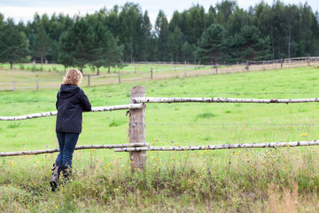 Woman enjoying rural view, standing her back to camera, keeping hands on wooden fence