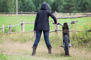 Rear view at female dressed black raincoat with hood on head standing with dirt motorcycle on countryside road