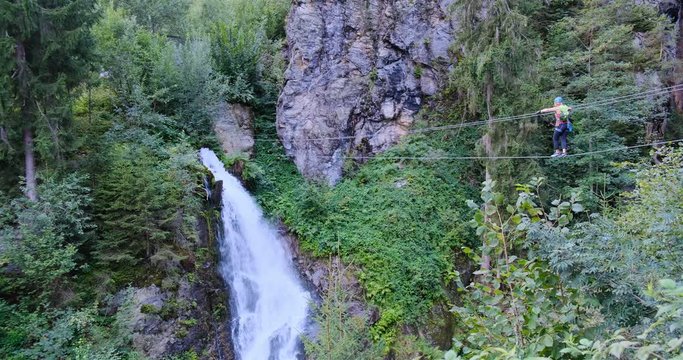 Woman advancing very carefully on a long via ferrata wire bridge towards the impressive, epic Talbach waterfall in Zillertal valley, Austria.