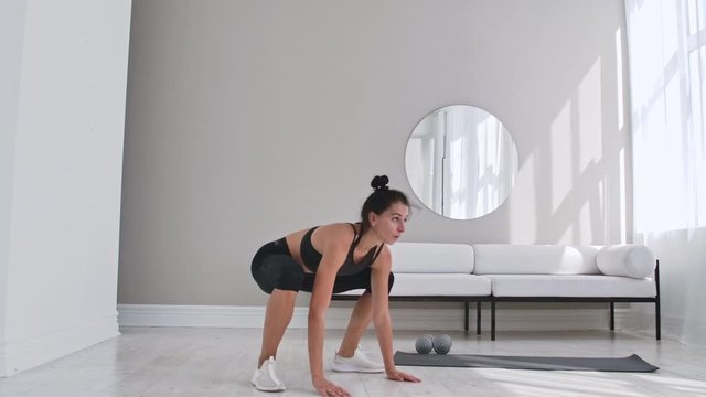 Beautiful slim woman in thirties with fair complexion does morning strenth and aerobic burpee exercises in modern airy room.