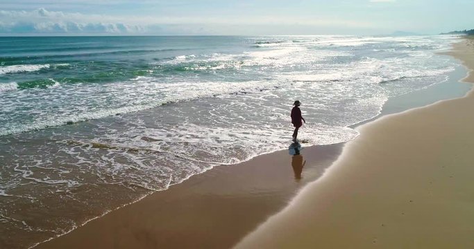 Drone y hermosa mujer con pamela paseando en soledadpor amplia playa de arena dorada 