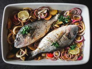 Two fresh doradas (gilt-head bream) with onion, tomatoes and herbs in white baking pan. Overhead shot.