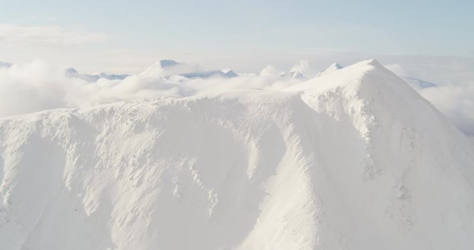 Aerial helicopter shot, following across snowy mountain peak, clouds and mountain range in the distance, drone footage