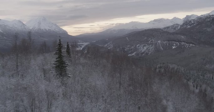 Aerial helicopter shot over mountain ridge with barren trees, reveals frozen river and mountains, drone footage
