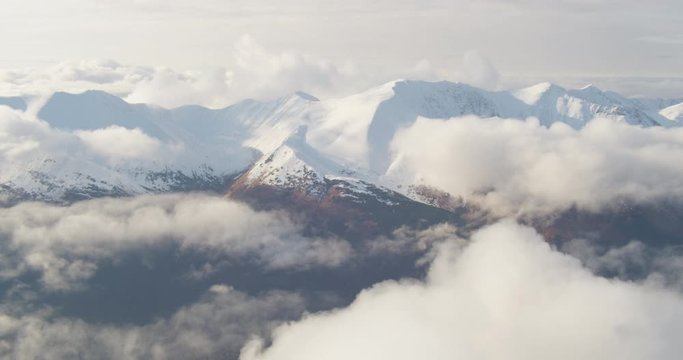 Aerial helicopter shot, tracking shot of idyllic snowy mountain range surrounded by clouds on a sunny day, drone footage