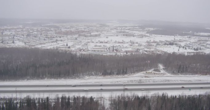 Aerial helicopter shot, wide tracking shot alongside highway in snowstorm, suburban town and mountains in the distance, drone footage