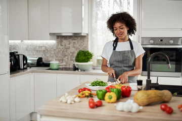 Gorgeous mixed race housewife in apron standing in kitchen and chopping mushrooms. On kitchen counter are all sorts of vegetables.