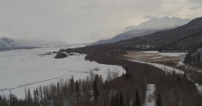  Aerial helicopter push past snowy highway adjecent to frozen river in snowy mountain valley, drone footage