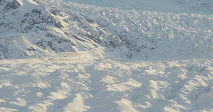  Aerial helicopter shot, track across details of rippling glacier at golden hour, zoom out to reveal tall snowy mountains, drone footage