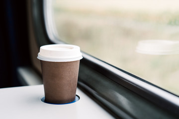 A paper coffee cup with a white plastic lid. Coffee to go on a table in the train overlooking a beautiful rural green landscape. Travel, lifestyle