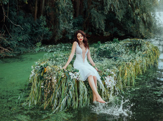 Fototapeta premium Cheerful woman in a white vintage outfit sits on a boat and splashes her bare feet in the water. wild background river with water lilies and green trees. Happy Feminine Princess deep neckline dress