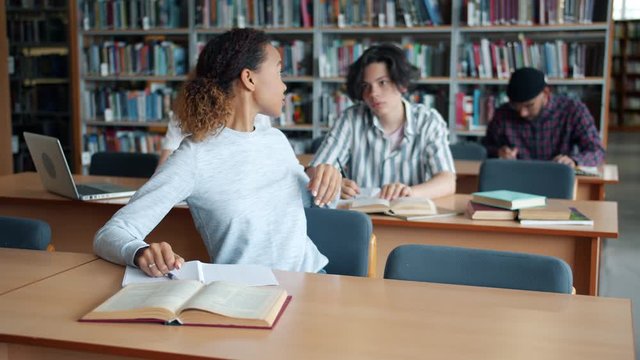 Cheerful African American girl is studying in university library reading taking book from male student sitting at desk. Lifestyle, education and literature concept.