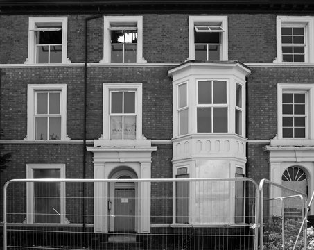 monochrome image of a row of abandoned dilapidated derelict houses in a street behind a metal fence in southport merseyside
