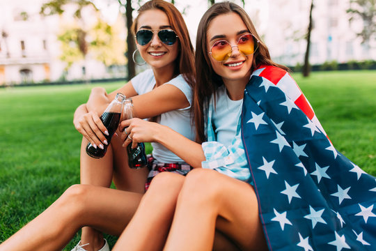 Two attractive young girls in sunglasses sitting in the Park on the lawn with the American flag, with a refreshing drink