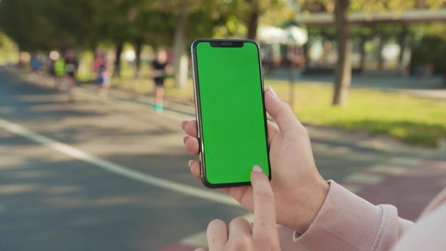 Slow motion woman hands holding and touching phone with green screen vertically in the street people running marathon on background internet technology business message close up