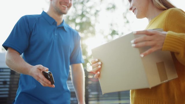 Beautiful Young Woman Meets Delivery Man who Gives Her Cardboard Box Package, She Signs Electronic Signature POD Device. Courier Delivering Parcel in the Suburban Neighborhood. Focus on the Box