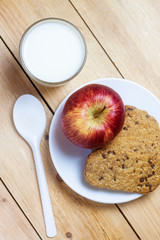 TOP VIEW OF BREAKFAST WITH MILK GLASS, COOKIES WITH CHOCOLATE CHIPS AND RED APPLE ON KITCHEN CLOTH AND WOODEN TABLE