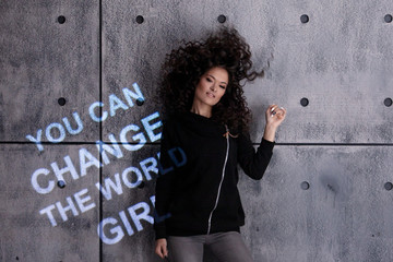 Curly girl dances against the background of gray wall onto which a multi colored motivational...