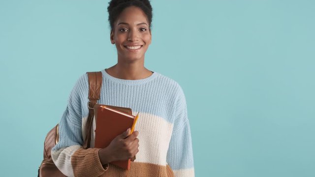 Pretty african american female student with backpack and notepad walking into shot and joyfully looking in camera over blue background