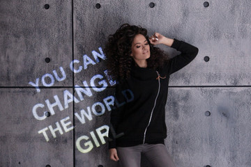 Fototapeta na wymiar Curly girl dances against the background of gray wall onto which a multi colored motivational inscription is projected.