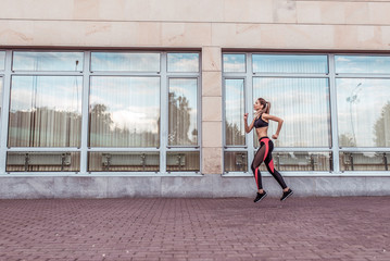 Fototapeta na wymiar Girl athlete runs background of glass windows, in summer in city on morning run, free space for text motivation fitness workout. Modern lifestyle of youth. Sportswear Leggings. Top. Sneakers.